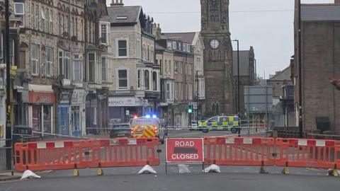 Road closed sign next to a police cordon. Police vehicles are parked next to the area. 