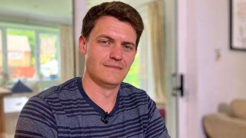 Dave in his home wearing a blue striped t-shirt, with short black hair, smiling at the camera.