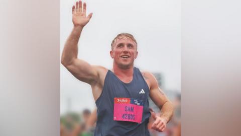 Sam Wealleans, who has blond hair and is wearing a dark blue running vest. He is smiling and waving to the crowd.