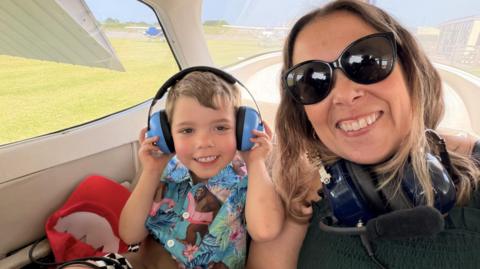 An image of Nicki Hale and her son sat in a small plane. They are both smiling at the camera. Nicki has a pair of headphones around her neck, and her son has them on his head. He looks about four years old and is wearing a brightly coloured shirt. 