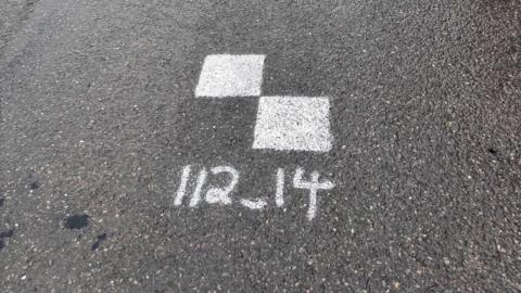 Two white squares painted on a road. Underneath is the number "112_14".