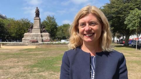A woman smiling with shoulder length blonde hair wearing a navy blazer and white and navy dotted top underneath. She is stood in a park with a large statue to the left of her and trees and parked cars to the right. The sky is blue with a few grey skies.