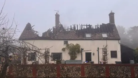 A house on a grey and misty morning. Its roof has been burnt in a fire and parts of the wooden structure can be seen jutting out from the tiles. Two chimney remain standing. 
