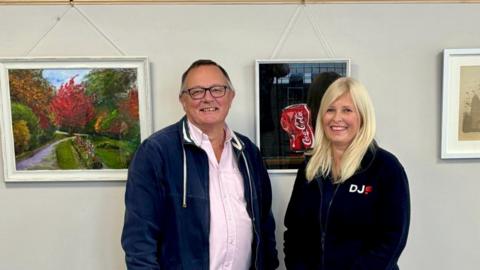 Keith Holdaway and a woman smiling for the camera in front of a wall of paintings. The woman wears a DJ Property branded top. One painting depicts a green space, while the other one is of a Coca Cola can Mr Holdaway is wearing a pink shirt and no tie with a casual blue jacket over the top. He also has a pair of glasses on