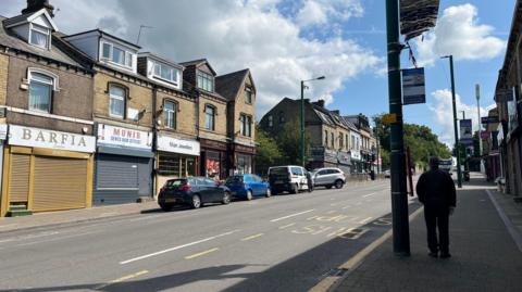 A row of shops on Leeds Road in Bradford, with a man stood waiting at a bus stop on the right side of the image.