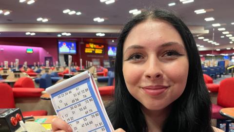 Lucy Lonnen, 18, has long black hair parted in the middle at the top of her forehead, she has a gold nose ring in her right nostril and is holding a bingo card up on the left hand side, behind her are red and blue sofa chairs in the bingo hall with large screens on walls in the far backgrou