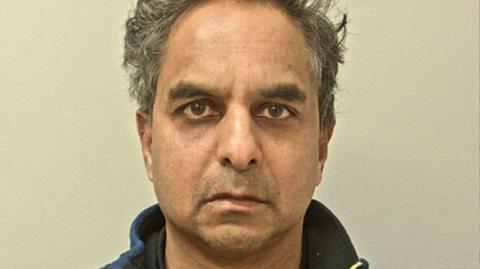 Police mugshot of Dr Amal Bose. He has short, unkempt dark hair that is greying and is looking directly at the camera, frowning slightly. He has brown eyes and bushy eyebrows.