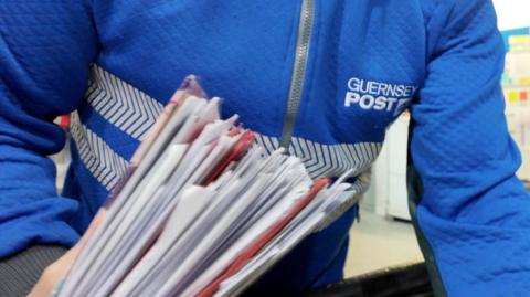 A close up picture of a Guernsey Post worker sorted through a large collection of letters and envelops. They are wearing a blue branded Guernsey Post jacket and are holding the assortment of envelopes and letter in their left hand. 