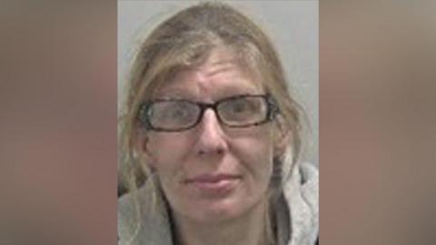 Katie has light brown hair tied up with strands framing her face. She wears black rectangular glasses and a black t-shirt with a grey hoodie