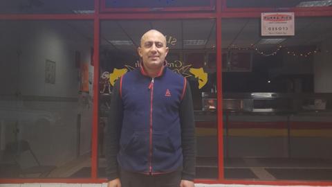 Jay Choganbaz standing outside his fish and chip shop in Lawrence Weston, Bristol, wearing a navy zip jumper with a shop window behind. There is a counter visible through the window and a pukka pies sign on the window