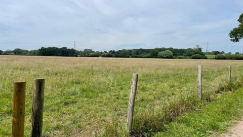 A large open field in Pewsham, Chippenham with trees and hedgerows in the background and fence posts with barbed wire in the foreground.