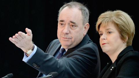Alex Salmond, left, wearing a dark suit and looking away from the camera, raising his left armd and hand in the air. Nicola Sturgeon, right, wearing a black top. Both are sitting against a black background.