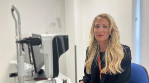 a woman with long blonde hair sitting at a desk with equipment for testing eyes 