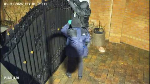 CCTV footage showing two men, wearing gloves and with their hoods raised, standing outside a large black wrought-iron gate. One man has both arms raised above the gate and out of frame. 