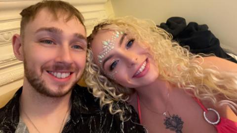 Billy with Katelen both smiling at the camera. He has short brown hair and is wearing an open-neck shirt. She has long wavy blond hair and has patterned beads on her head. She is wearing a pink halterneck top.