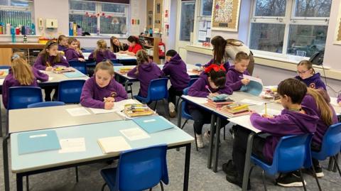 A classroom with about 19 children sat across five tables with books and pens. They are wearing a uniform of purple hoodies. Two teachers or assistants, at separate tables, are each talking to a youngster.