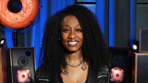 A woman with curly black hair smiling at the camera. She is wearing a silver necklace and a black leather jacket and behind her is sound equipment.