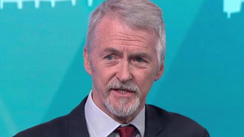 Huw sits in the politics wales studio in a shot taken from the programme. He is wearing a dark suit and red die with a remembrance poppy on his lapel He has short grey hair and a grey goatee style beard.