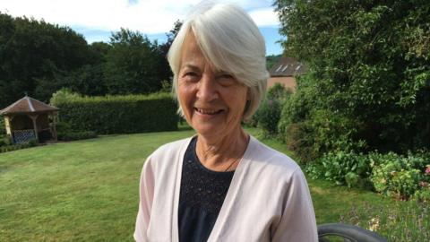 Pat Pope smiles at the camera in a garden wearing a pink cardigan and black top