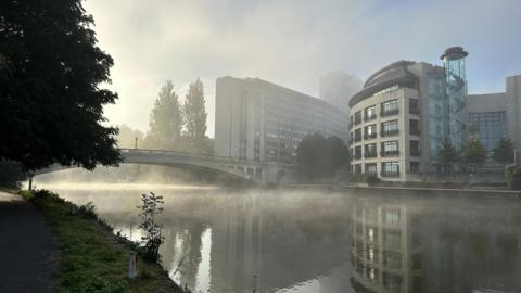 Mist rolls over a river in a built-up suburban area. A bridge crosses the river with apartment blocks on one side. Trees can be seen on the other side under a murky sky, although part of it is blue.