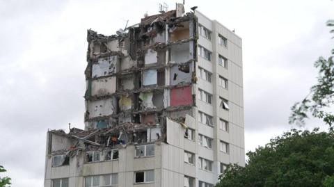 A tower block that is part demolished. Rows of flats have been taken out. It is standing against a grey background.