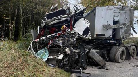 Wreckage of a lorry cab which appears to have been crushed in a crash with a trailer behind which appears to have partially escaped the collision with trees on each side of a lane and the remains of the cab resting on a grassy bank.