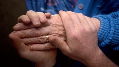 A pair of hands wrapped around the hand of an elderly person who is wearing a gold wedding band and a blue knitted cardigan.