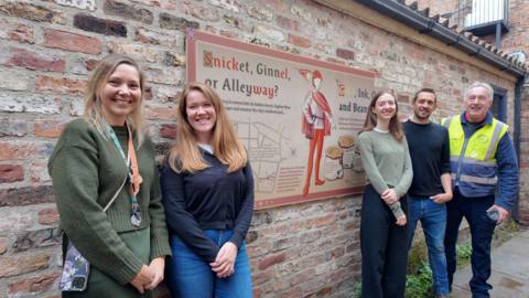 Three women and two men stand in a snickelway, lined with a brick wall. On the wall is artwork with the title 'snicket, ginnel or alleyway?' and displays a picture of a medieval man.