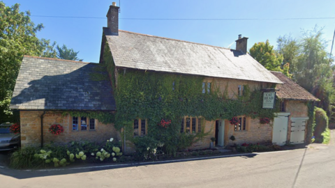 A light stone pub building, much of which is covered in ivy. It has a pistachio coloured sign with the pub's name on it. There are hydrangeas growing in front of the pub and it is a sunny day.