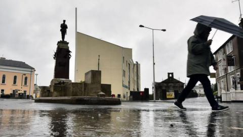 The cenotaph in Enniskillen on a rainy day, a passer by is to the right of shot with an umbrella and grey coat.