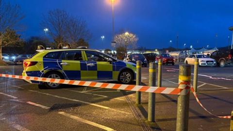 Red and white striped tape cordons off part of the car park outside a Tesco supermarket. A police car is parked within the cordon. It is dark. There are other cars in the background.