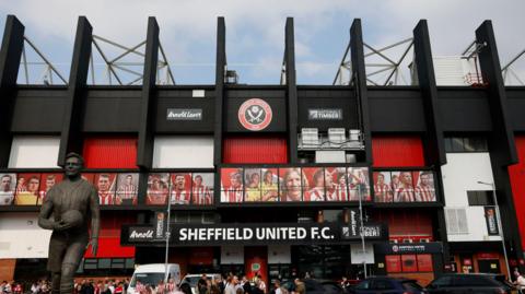 Fans can be seen in front of the entrance to Bramall Lane, which features a sign that reads "Sheffield United FC". A bronze statue of a man can be seen on the left. The stadium's colour scheme is white and red.