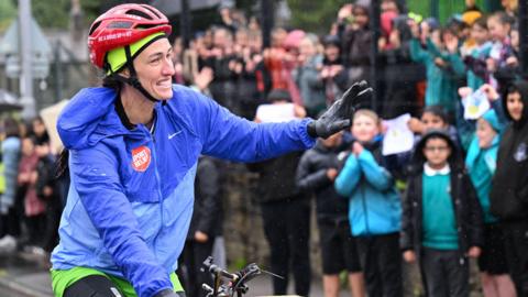 Jill Scott is on her bike but you can only see the handlebars. She is wearing black leggings, green shorts, a light blue jacket with the Sport Relief logo which is white letters on a red background. She is wearing a red cycle helmet with a fluorescent green hat underneath. She has one hand waving to the crowd and is wearing a black glove. Supporters can be seen lining the route behind her but they are blurred