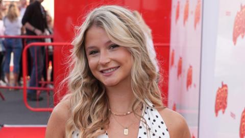 Mollie Pearce, a young woman with long, curly blonde hair, smiles at the camera at a red carpet event. She is wearing a black and white polkadot halterneck top.