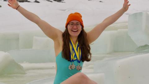 Woman (Erin Medcalf) with long brown hair with an orange hat and glasses on smiles brightly holding her hands up in celebration as she sits on and surrounded by ice blocks in a turquoise blue swim suit. She had four medals around her neck.