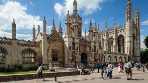 King's College in Cambridge on a sunny day. There are people walking on the pavement and public road in front of the gate and its chapel. 