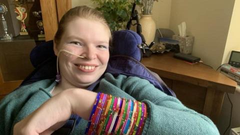 Lucinda Ritchie is smiling, seated in a supportive chair raising an arm covered with multiple colourful beaded bangles. She has an oxygen tube. She is wearing a textured, long‑sleeved top. In the background are a wooden cabinet with trophies, a desk with assorted items, and decorative plants. 