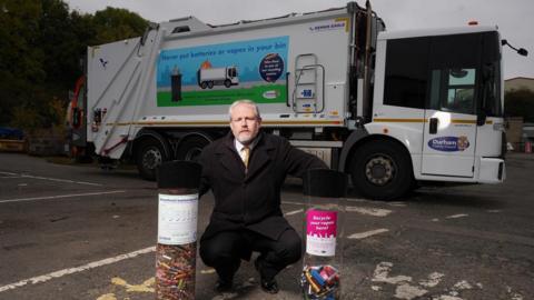 Mark Wilkes, Durham County Council's member for climate change. He has short grey hair, a short grey beard and is wearing a black coat over a dark suit with white shirt and yellow tie. He is squatting with his hands resting on two waist-high, tubular clear plastic recycle bins. A large white bin lorry is parked behind him.