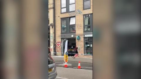 A black BMW that has crashed into a pizza restaurant. There is police tape and traffic cones cordoning off the scene.