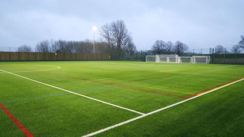 A green 3G pitch with some lit floodlights. There is a brown fence running down the side. There are portable football goals in the background.