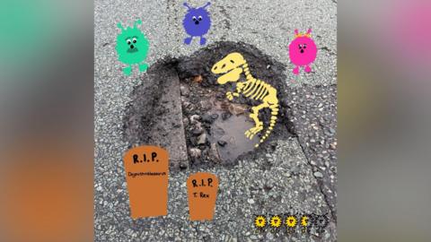 A photo of a pothole in a road that has drawings on top of it. The drawings depict a dinosaur skeleton in the pothole, with two grave stones below it. Above the pothole are three fuzzy creatures - one pink, one blue and one green