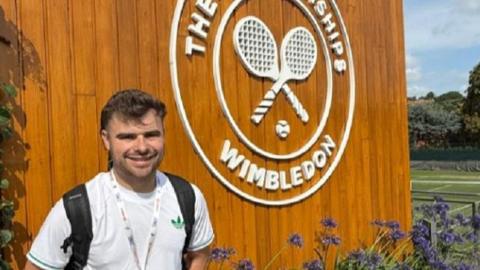 Matthew Brough stood in front of the Wimbledon sign on a brown board, with tennis courts and purple flowers in the background. He is wearing a white t-shirt with a green Adidas logo and has a backpack on, with styled, short brown hair. 