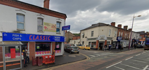 Google street view of a road junction. A fish and chip kebab house can be seen on the left, with a blue sign and a bus shelter outside. On the opposite corner is a south Asian sweet shop with a grey sign. Pedestrians can be seen walking on the pavement and other shops in the distance occupying the ground floors of terraced buildings.