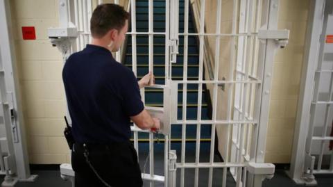 Rearview of a prison officer locking a white barred gate, with a staircase behind it
