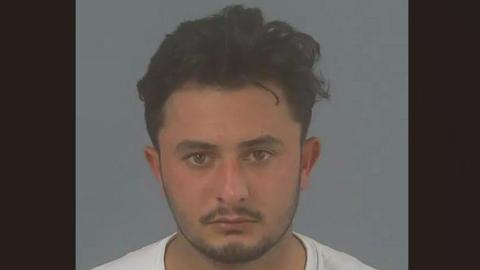 Musafar Hotak, a man with dark hair and a light beard, is pictured facing the camera in a police mugshot. He is wearing a white T-shirt.