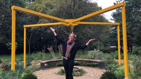 Kate Smithson standing in a garden beneath a structure of yellow iron beams