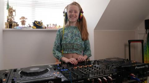 A young girl with long blonde hair is in her house smiling and stood behind some large black DJ decks full of dials and lights.