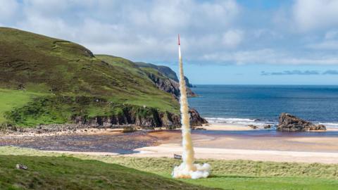 Rocket launches from grass-covered land near a beach by the sea