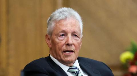 Peter Robinson. He has short white hair, wearing a navy blazer, white shirt and green and navy tie.