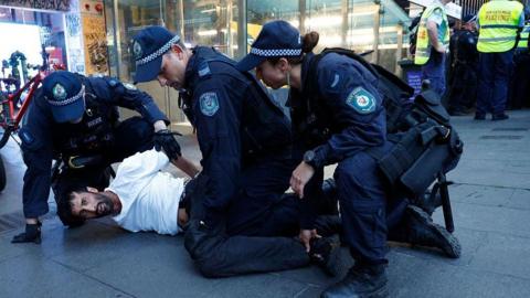 A man wearing a white t-shirt is held on the ground by three police officers.